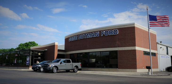 Exterior of 'Gene Butman Ford' dealership with a brick facade, large windows, and two cars parked outside. 
