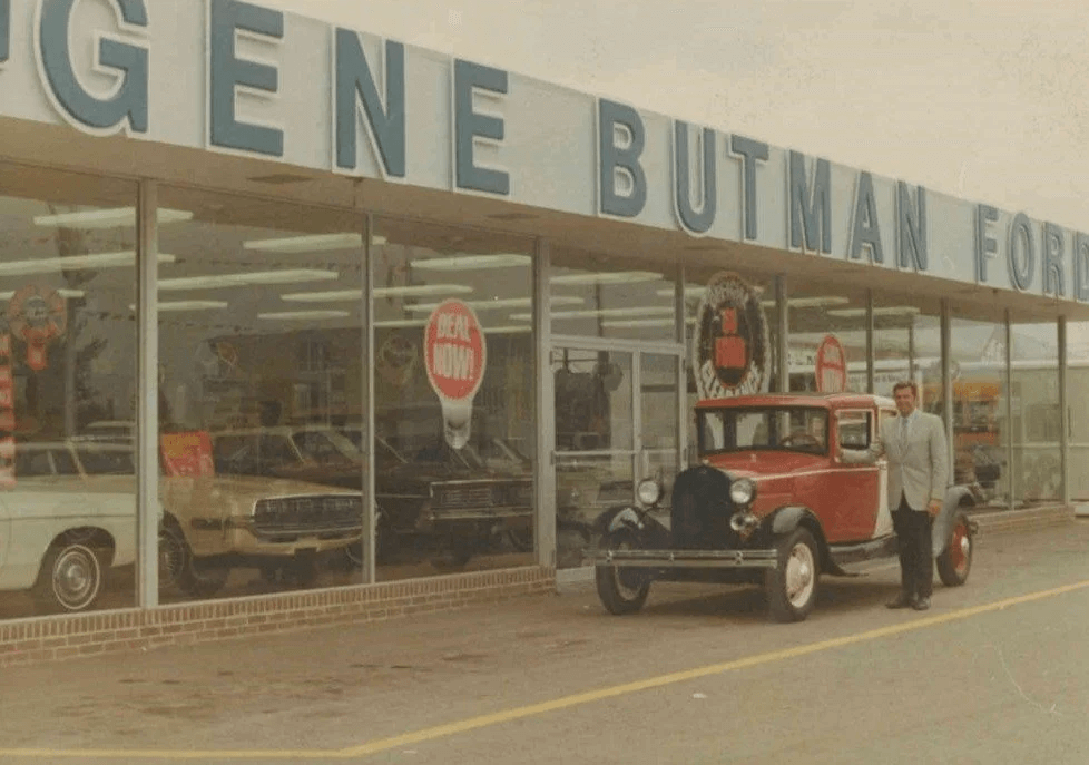 A person, in a suit, stands next to a red Model A Ford with a dealership in the background.
